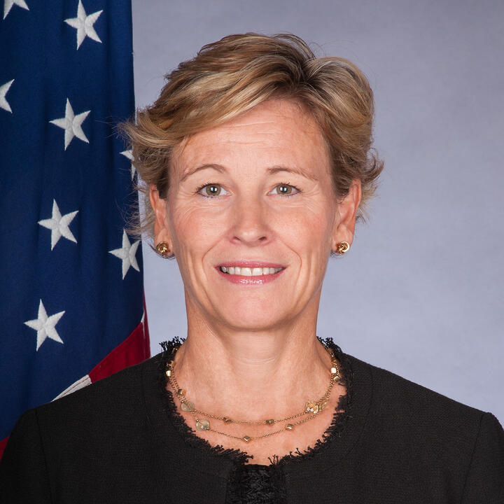 Photo of Jennifer Haskell, a blonde short-haired woman with hazel eyes standing in front of a U.S. Flag. She is wearing a black blazer and a gold necklace with gemstones.